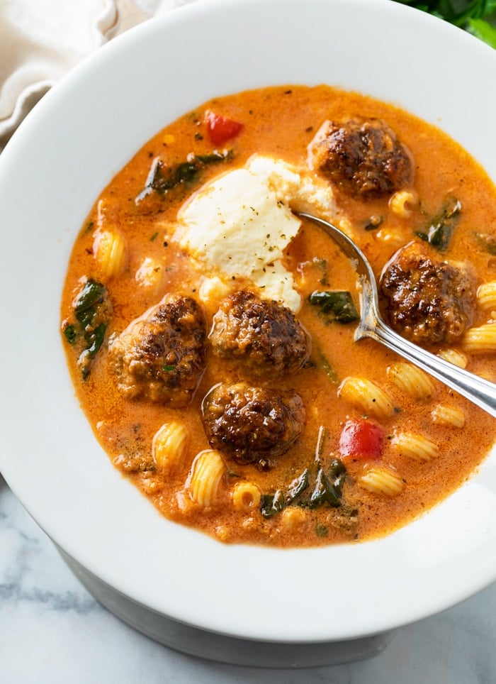 A white bowl with meatball soup topped with ricotta cheese with a spoon on the side.