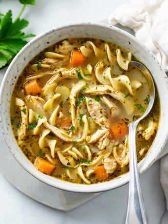 A bowl of chicken noodle soup with a spoon in the bowl and parsley.