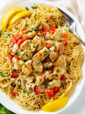 A white platter with Chicken Scampi topped with diced tomatoes, parsley, and slices of lemon.