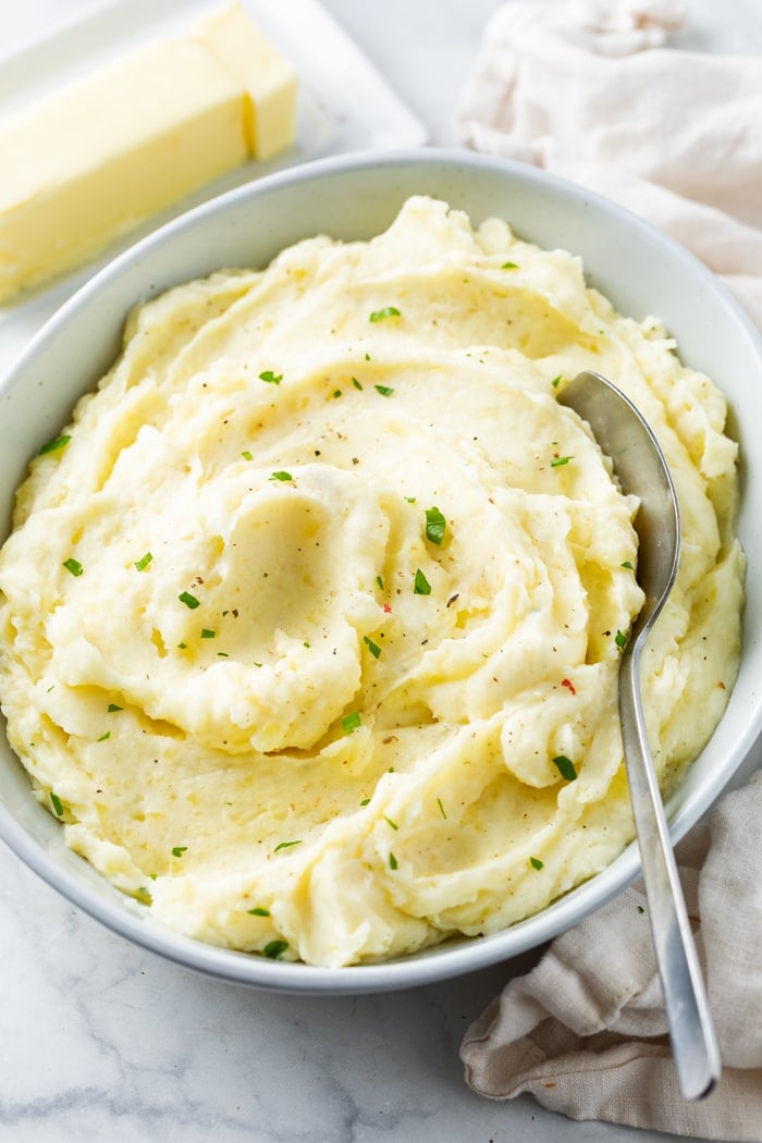 A white bowl of creamy homemade mashed potatoes topped with parsley with butter in the background.