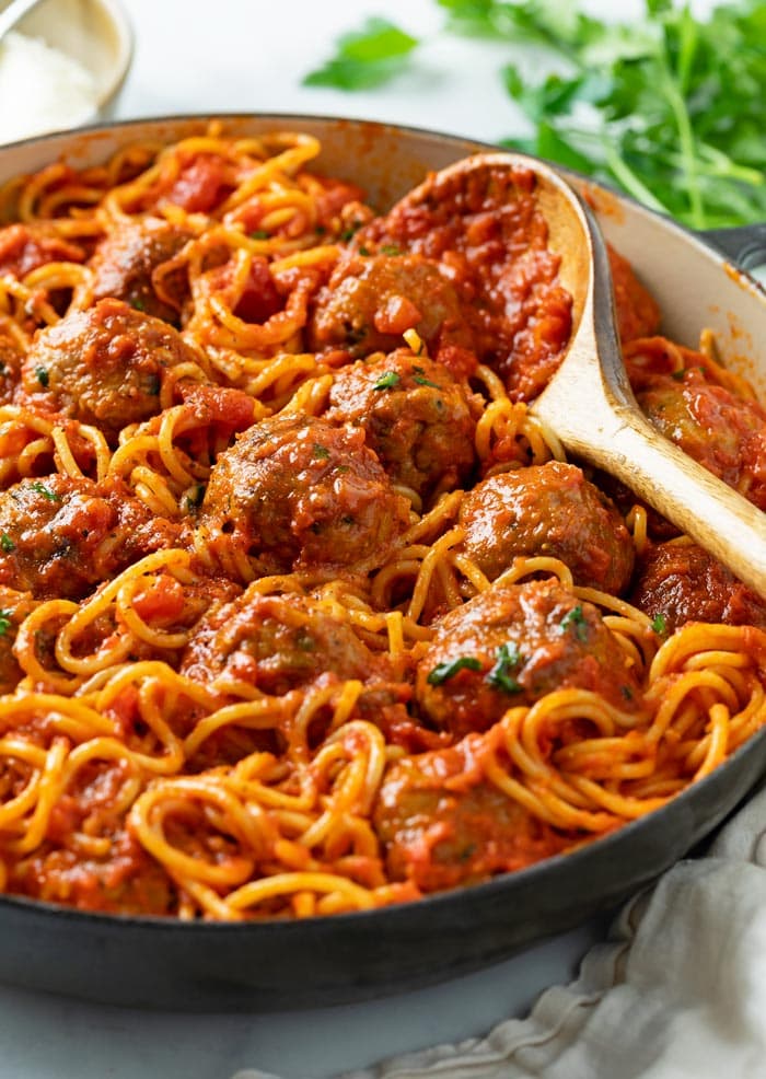 A pot of Spaghetti and Meatballs with a wooden spoon on the side and parsley in the background.