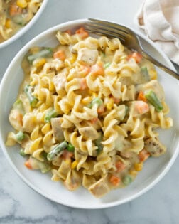 A white plate with creamy chicken and noodles with vegetables and a fork on the side.