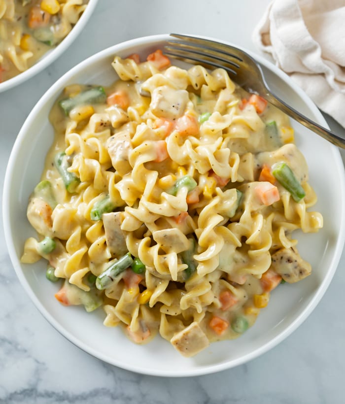 A white plate with creamy chicken and noodles with vegetables and a fork on the side.