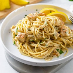 A white plate of creamy lemon chicken pasta with lemon slices and fresh parsley.