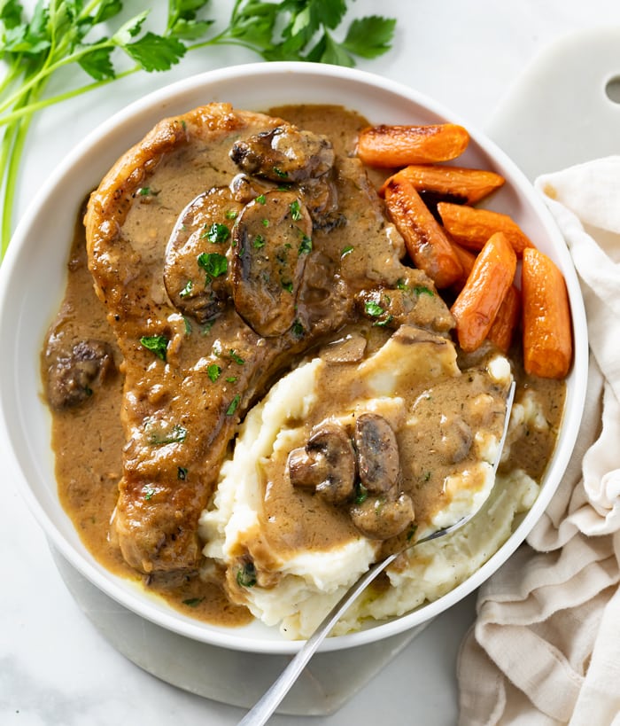 A white plate with Mushroom Pork Chops on it with mashed potatoes and carrots.