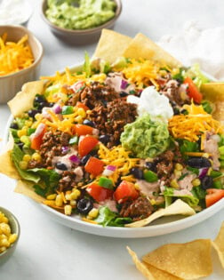 A white plate filled with Taco Salad topped with ground beef, avocado, sour cream, tortilla chips, and vegetables.