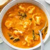 A bowl of creamy Tomato Tortellini Soup with spinach in it and a spoon on the side.