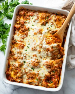 A white casserole dish with Baked Ravioli with a wooden spoon in it.