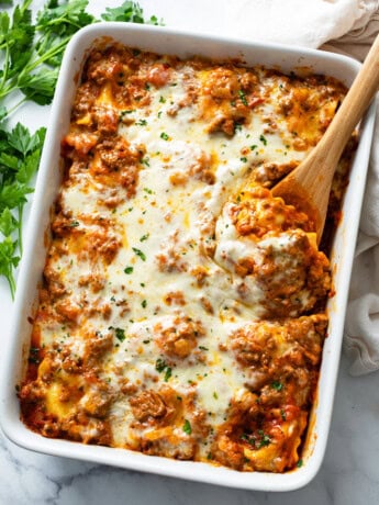 A white casserole dish with Baked Ravioli with a wooden spoon in it.