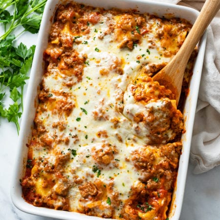 A white casserole dish with Baked Ravioli with a wooden spoon in it.