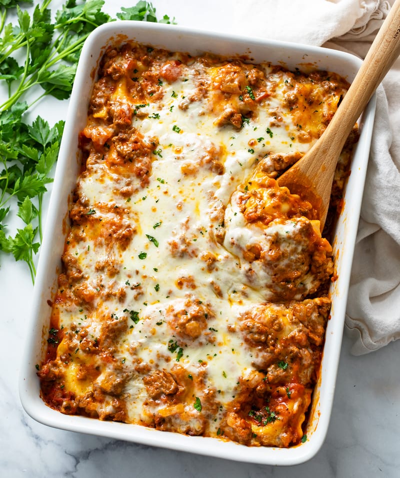 A white casserole dish with Baked Ravioli with a wooden spoon in it.