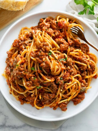 Spaghetti topped with Meat Sauce on a white plate with a fork and parsley on top.