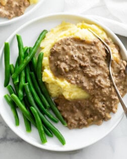 A pile of potatoes topped with Hamburger Gravy on a plate with green beans.