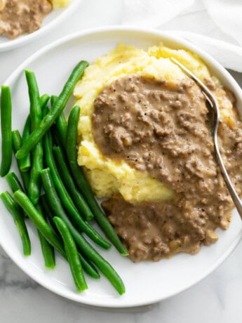 A pile of potatoes topped with Hamburger Gravy on a plate with green beans.