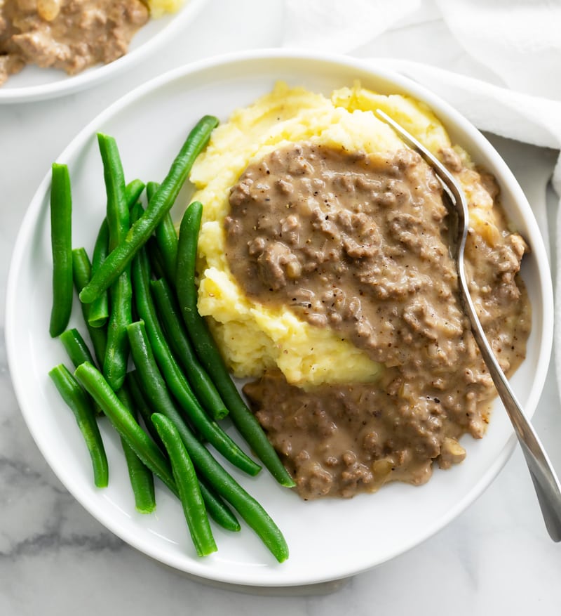 A pile of potatoes topped with Hamburger Gravy on a plate with green beans.