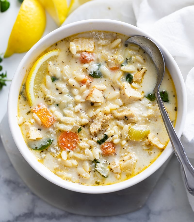 A white bowl filled with Lemon Chicken Orzo Soup with a lemon wedge and a spoon.