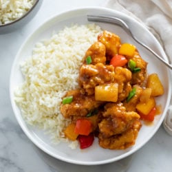 A white plate with Sweet and Sour Chicken with a side of white rice and green onions on top.