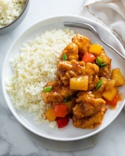 A white plate with Sweet and Sour Chicken with a side of white rice and green onions on top.