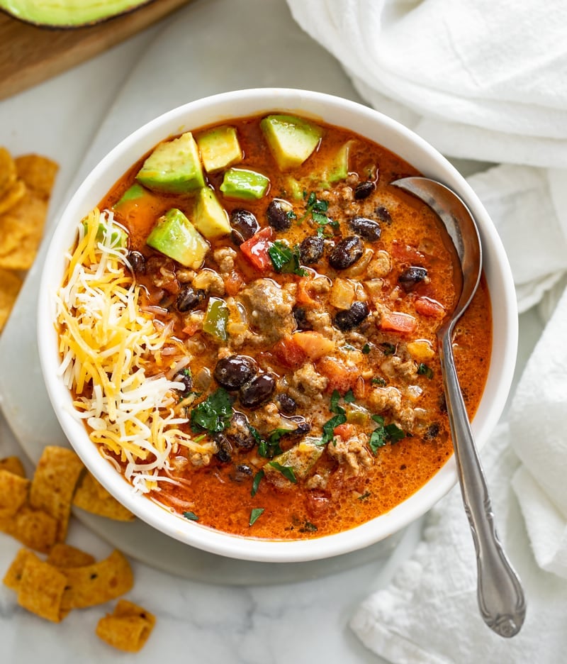 A white bowl filled with taco soup garnished with diced avocado and shredded cheese.