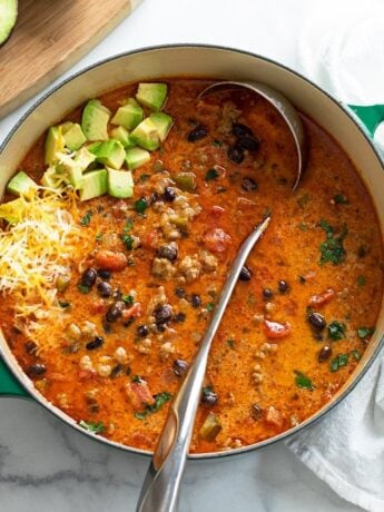 A soup pot filled with Taco Soup with a ladle in it and avocados and cheese on top.