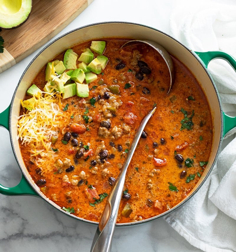 A soup pot filled with Taco Soup with a ladle in it and avocados and cheese on top.