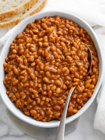 Baked Beans in a white bowl with a spoon and bread on the side.