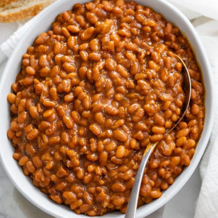 Baked Beans in a white bowl with a spoon and bread on the side.