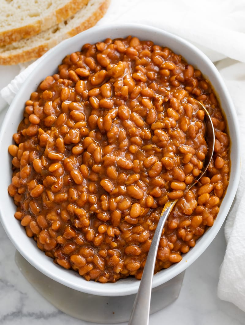 Baked Beans in a white bowl with a spoon and bread on the side.