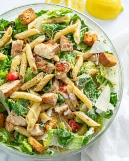 A glass bowl filled with Chicken Caesar Pasta Salad with Lemon wedges on the side.