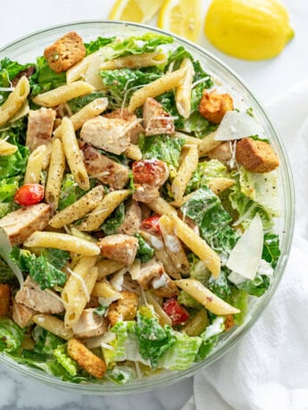 A glass bowl filled with Chicken Caesar Pasta Salad with Lemon wedges on the side.