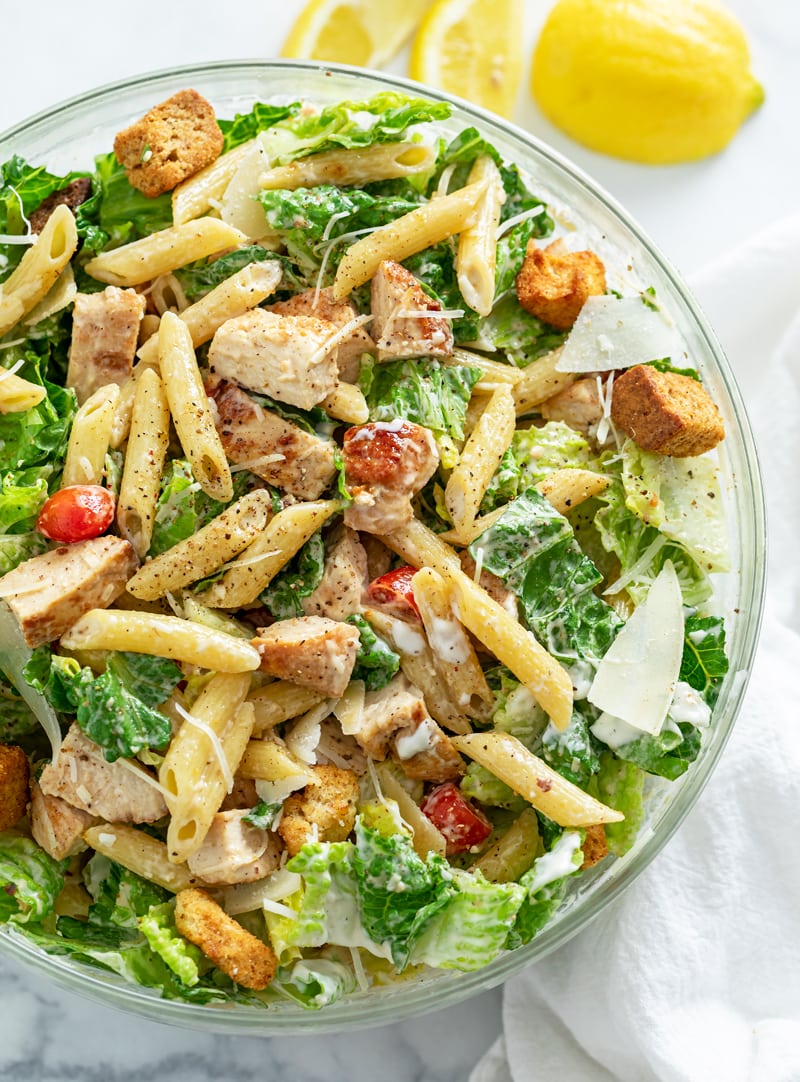A glass bowl filled with Chicken Caesar Pasta Salad with Lemon wedges on the side.