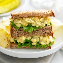 A white plate with a stack of egg salad sandwiches with chips in the back.