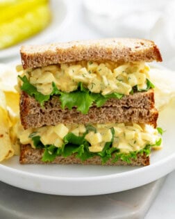 A white plate with a stack of egg salad sandwiches with chips in the back.