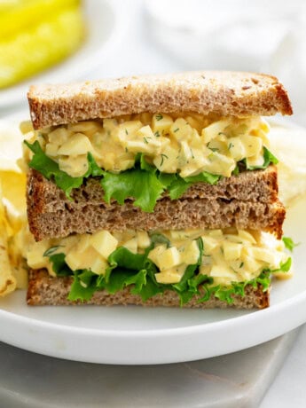 A white plate with a stack of egg salad sandwiches with chips in the back.