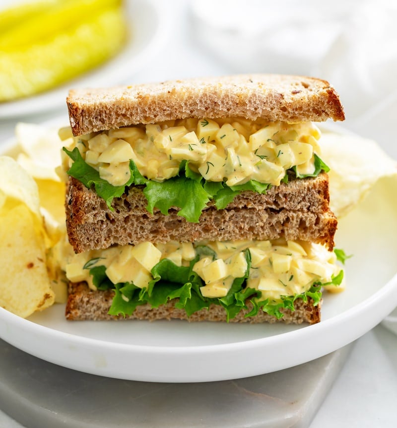 A white plate with a stack of egg salad sandwiches with chips in the back.