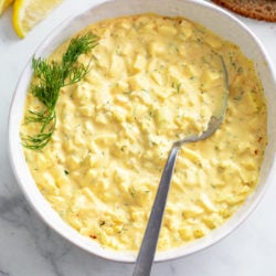 A white bowl filled with egg salad with fresh dill on the side and lemon wedges.
