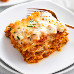 A white plate with Lasagna on it with a fork in the background.