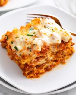 A white plate with Lasagna on it with a fork in the background.