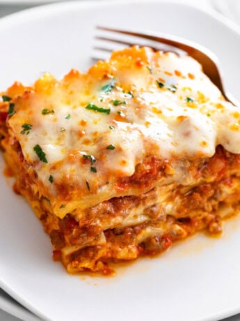 A white plate with Lasagna on it with a fork in the background.