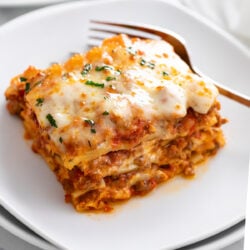 A slice of Lasagna on a white plate with parsley on top and a fork in the back.