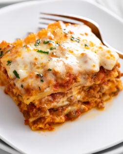 A slice of Lasagna on a white plate with parsley on top and a fork in the back.