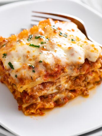 A slice of Lasagna on a white plate with parsley on top and a fork in the back.