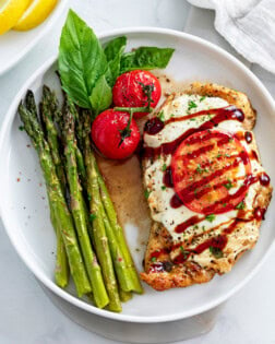 A white plate with Caprese Chicken next to roasted asparagus and fresh tomatoes and basil.