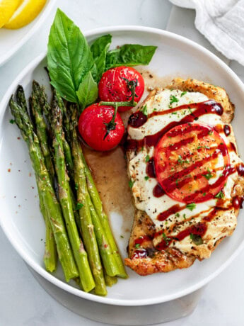 A white plate with Caprese Chicken next to roasted asparagus and fresh tomatoes and basil.