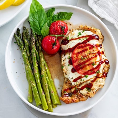 A white plate with Caprese Chicken next to roasted asparagus and fresh tomatoes and basil.
