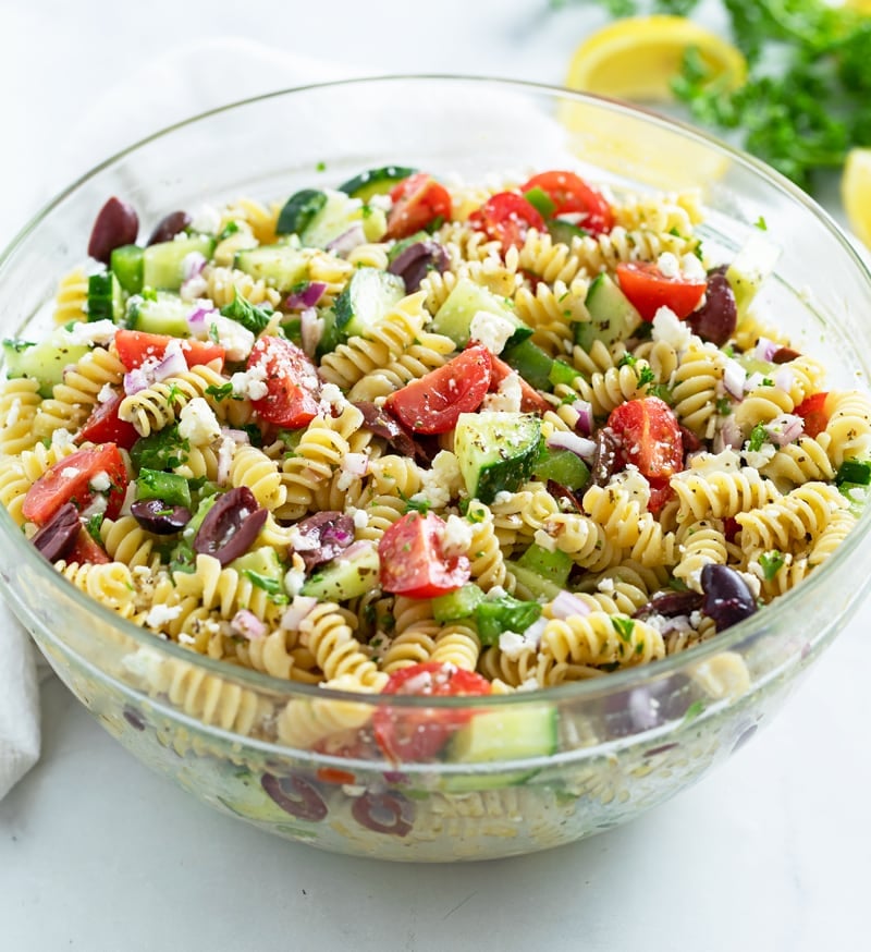 A glass bowl filled with Greek Pasta Salad with parsley and lemons in the background.
