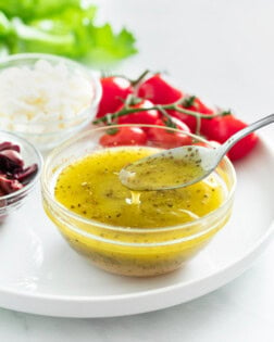 A spoon taking Greek Salad Dressing from a glass cup on a white plate.