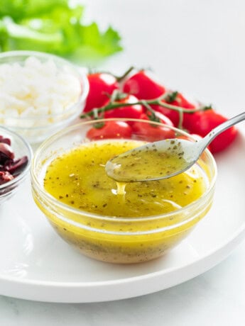 A spoon taking Greek Salad Dressing from a glass cup on a white plate.