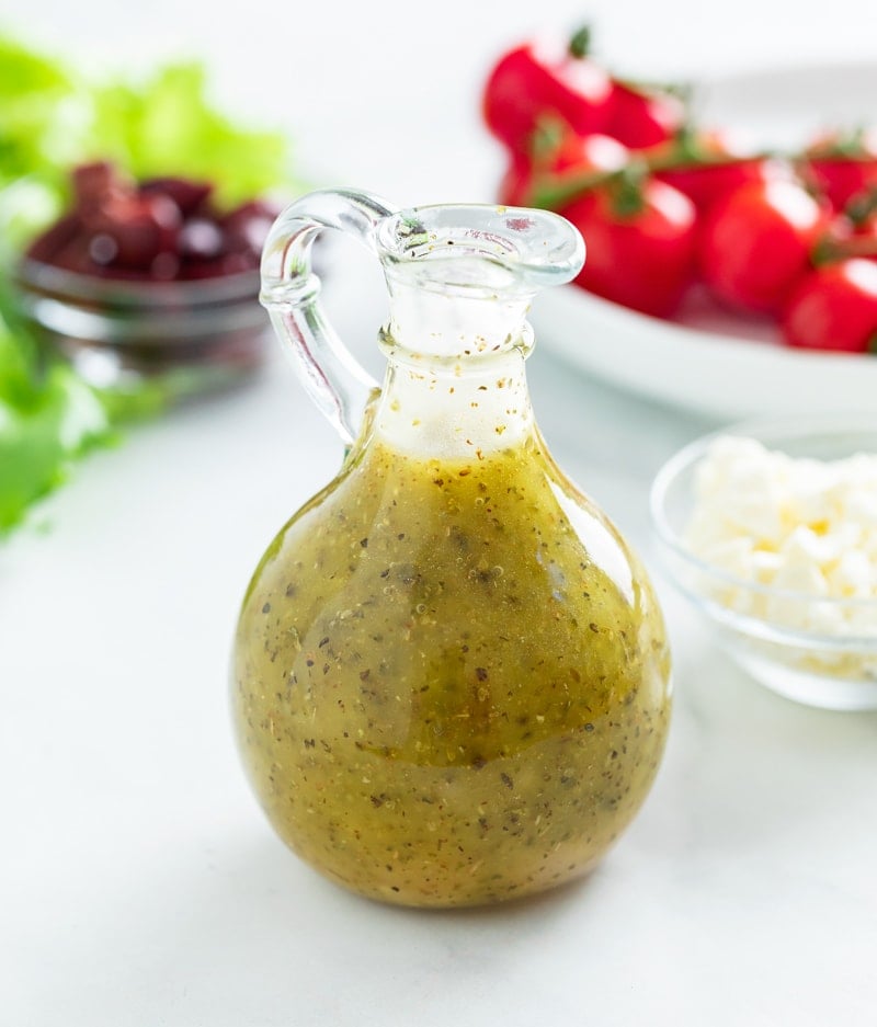 A bottle of Greek Salad Dressing with tomatoes, olives, and lettuce in the background.