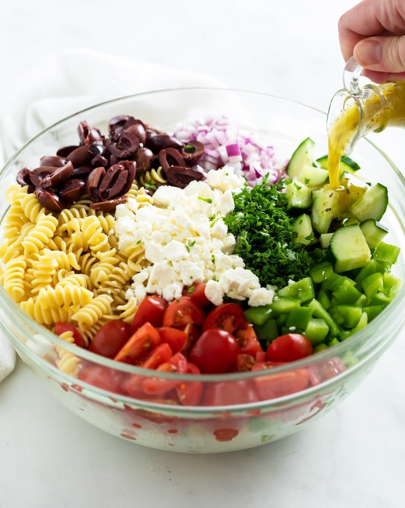 A glass bowl filled with ingredients to make Greek Pasta Salad.